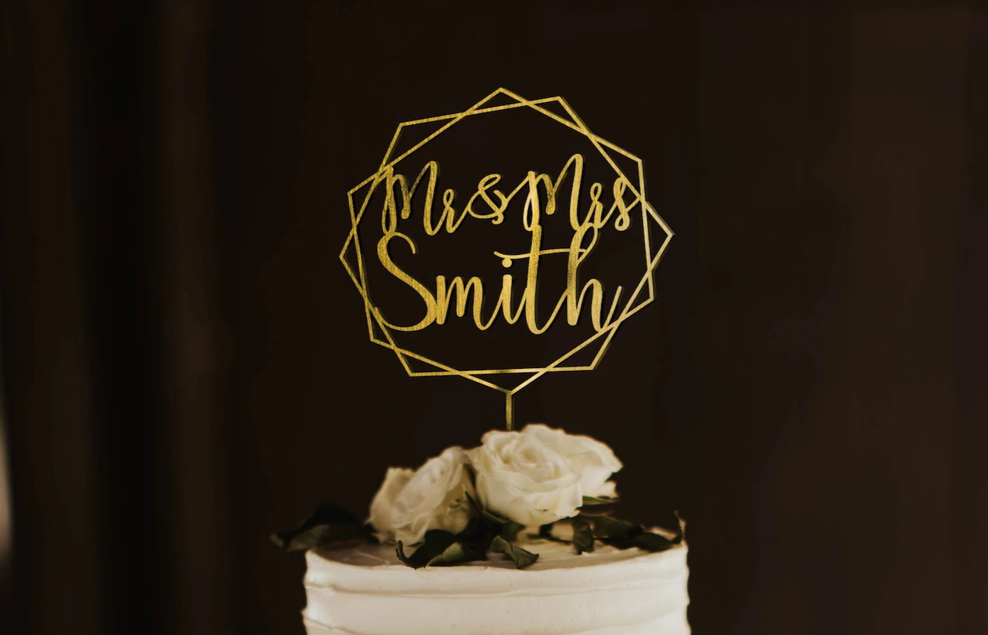 calligraphy personalized golden hexagon wreath style cake topper made of wood on top of the white simple wedding cake.
