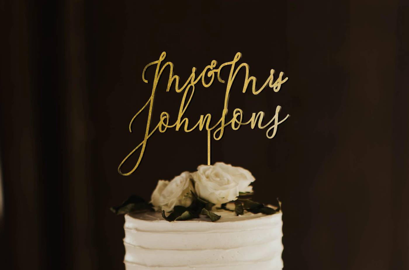 thin calligraphy personalized golden cake topper made of wood on top of the white simple wedding cake.
