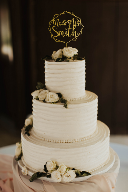 calligraphy personalized golden hexagon wreath style cake topper made of wood on top of the white simple wedding cake. 
