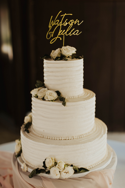 thin calligraphy personalized golden cake topper made of wood on top of the white simple wedding cake. 